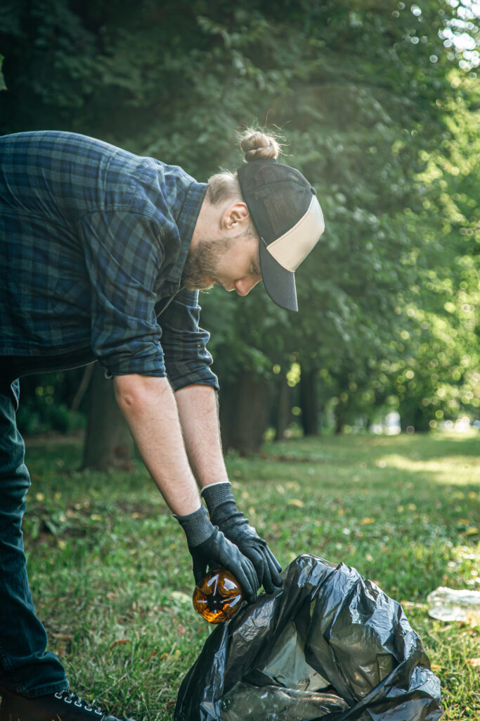 A young man with a trash bag in the forest cleans up plastic bottles.